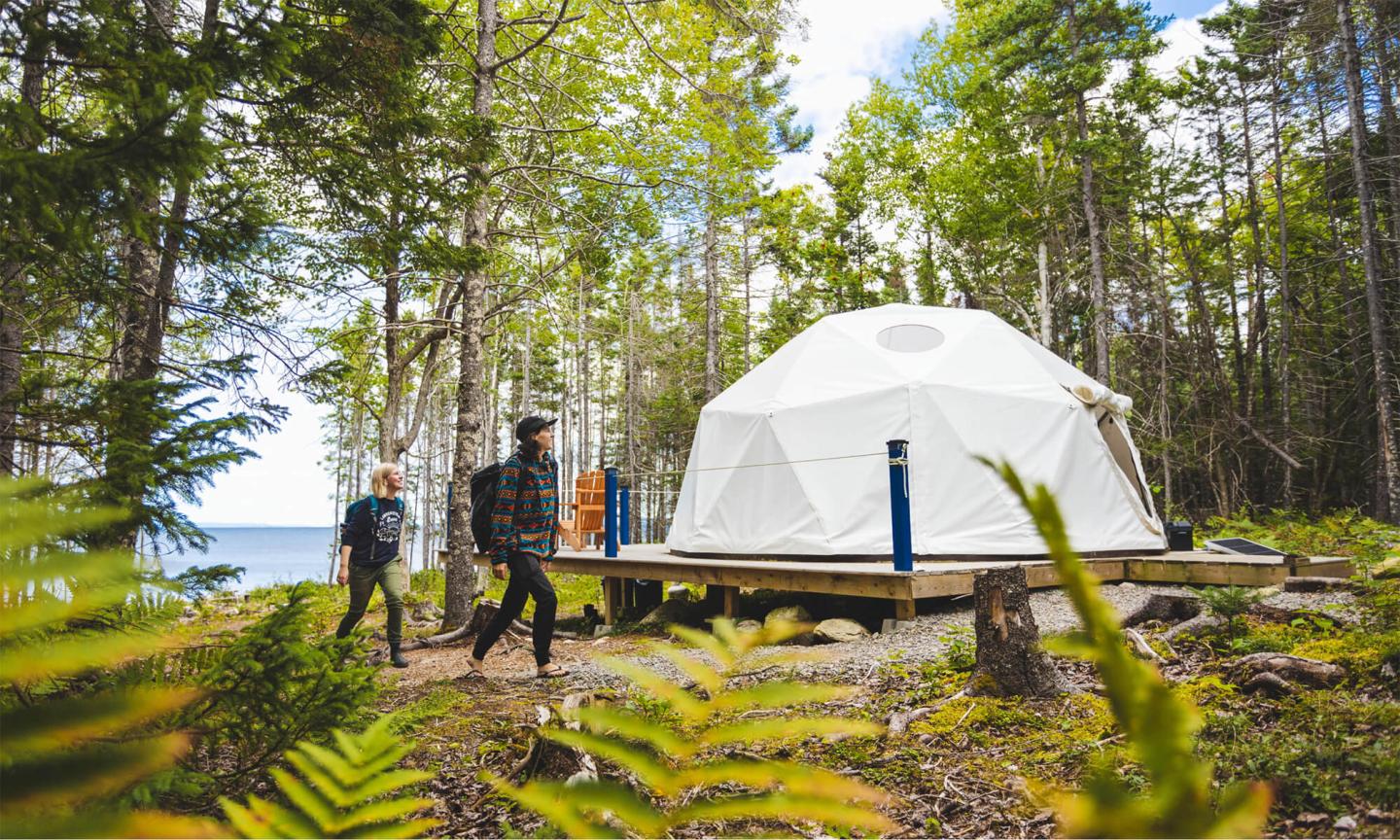 Two hikers walking toward a modern white glamping dome in a sunny forest.